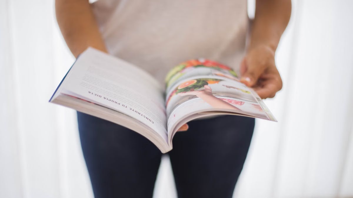 Close-up of a woman holding an open cookbook with colorful food images.